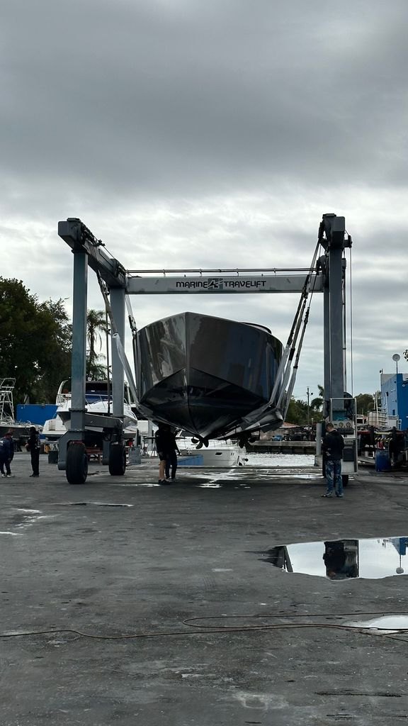 A boat suspended bow on in the straps of a Marine Travelift at a South Florida yard, the hull bottom exposed and yard crew alongside